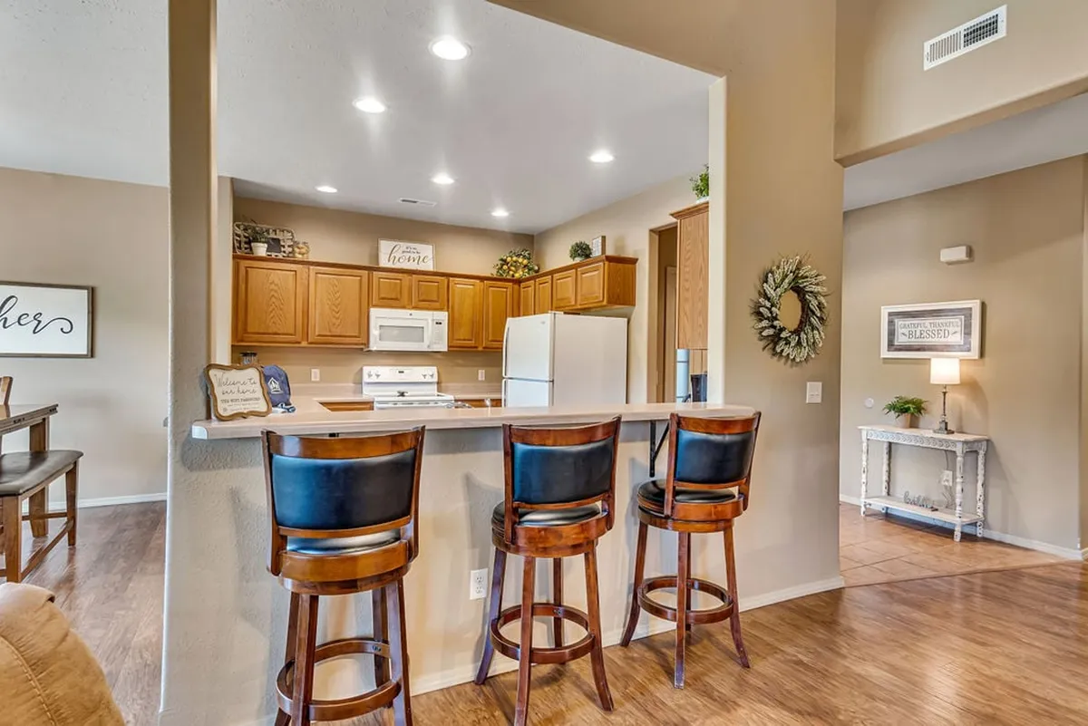 A modern kitchen with a large island, three bar stools, wooden cabinetry, decorative plants, and framed wall art. The space is designed for cooking, socializing, and family gatherings.