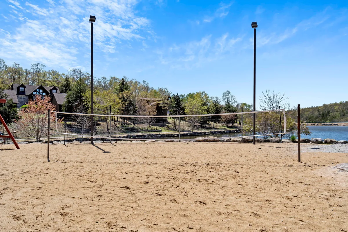 A dynamic outdoor setting featuring a sand volleyball court, a nearby playground, a lake, and distant buildings nestled among trees. The image emphasizes a wide array of activities available within a lush, natural setting.