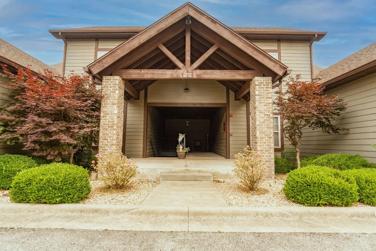 The exterior entrance of a resort building features inviting wooden beams, brick pillars, and landscaped shrubs. The scene suggests a welcoming arrival and conveys the ambiance of a peaceful, resort-style haven awaiting guests after a day out.