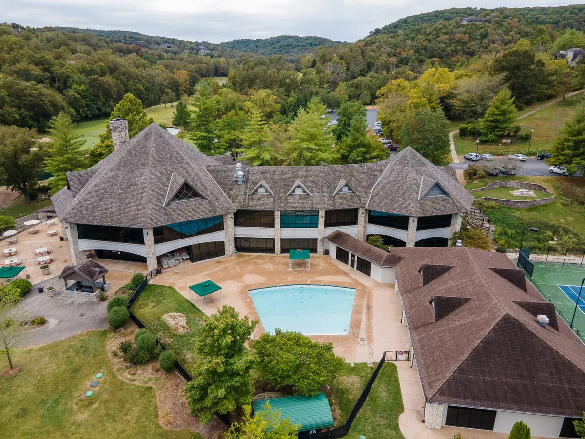 A vibrant exterior scene showing a sparkling swimming pool surrounded by lounge chairs, landscaped gardens, tennis court, and scenic mountain views in the background. The image radiates a lively, communal atmosphere perfect for family fun and outdoor activities.