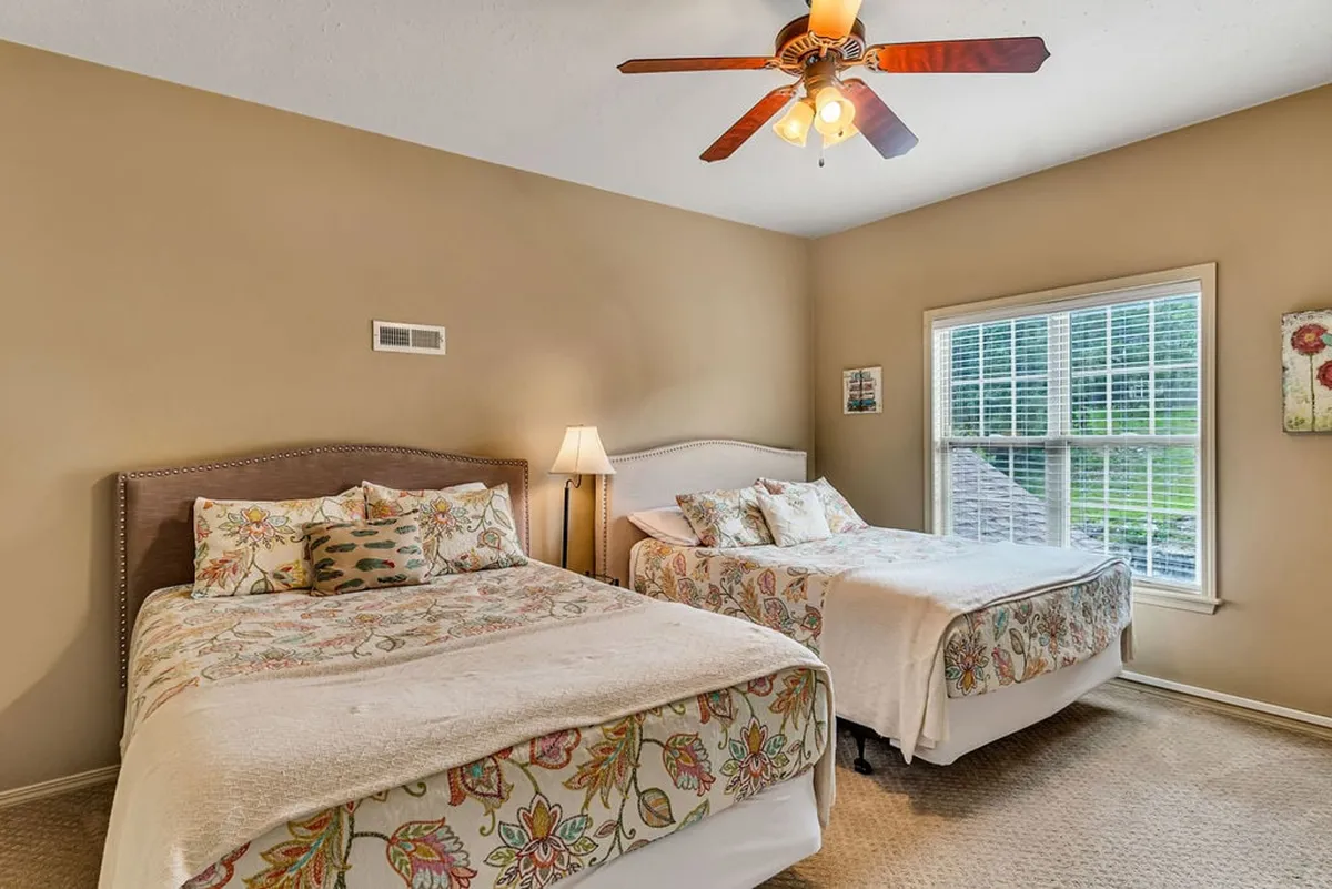 A cozy upstairs bedroom with two beds covered in floral bedding, a window allowing in natural light, and a ceiling fan overhead. The space feels inviting for both siblings and friends to enjoy sleepovers.