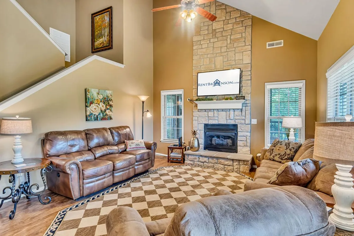 The image showcases a warm and inviting living room, featuring a striking stone fireplace as the centerpiece, surrounded by plush leather sofas and tasteful decorative artwork. Soft lighting from elegant lamps creates an atmosphere perfect for relaxation, conversation, or unwinding after a day on the golf course.