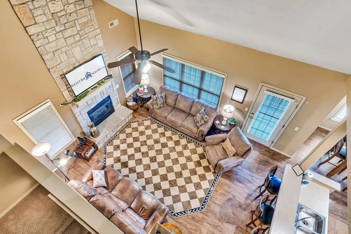 A cozy living room featuring a large stone fireplace, a spacious sectional sofa, a decorative area rug, and a flat-screen TV. Large windows bring in natural light, creating a warm and inviting atmosphere.
