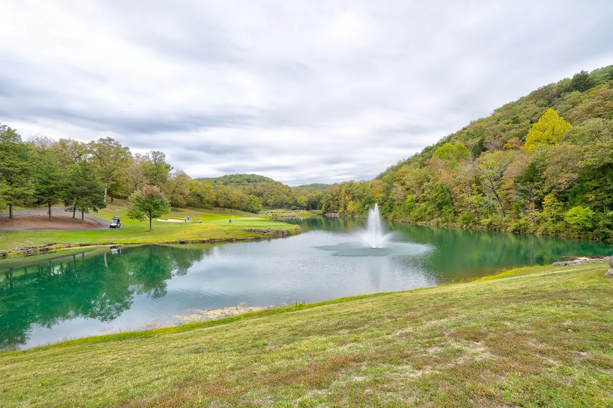 A serene lake with a central fountain, surrounded by rolling green grass, mature trees, and gentle hills in the distance. The peaceful water and natural landscape evoke a sense of tranquility and offer a perfect setting for fishing, picnicking, walking, or simply relaxing by the water.