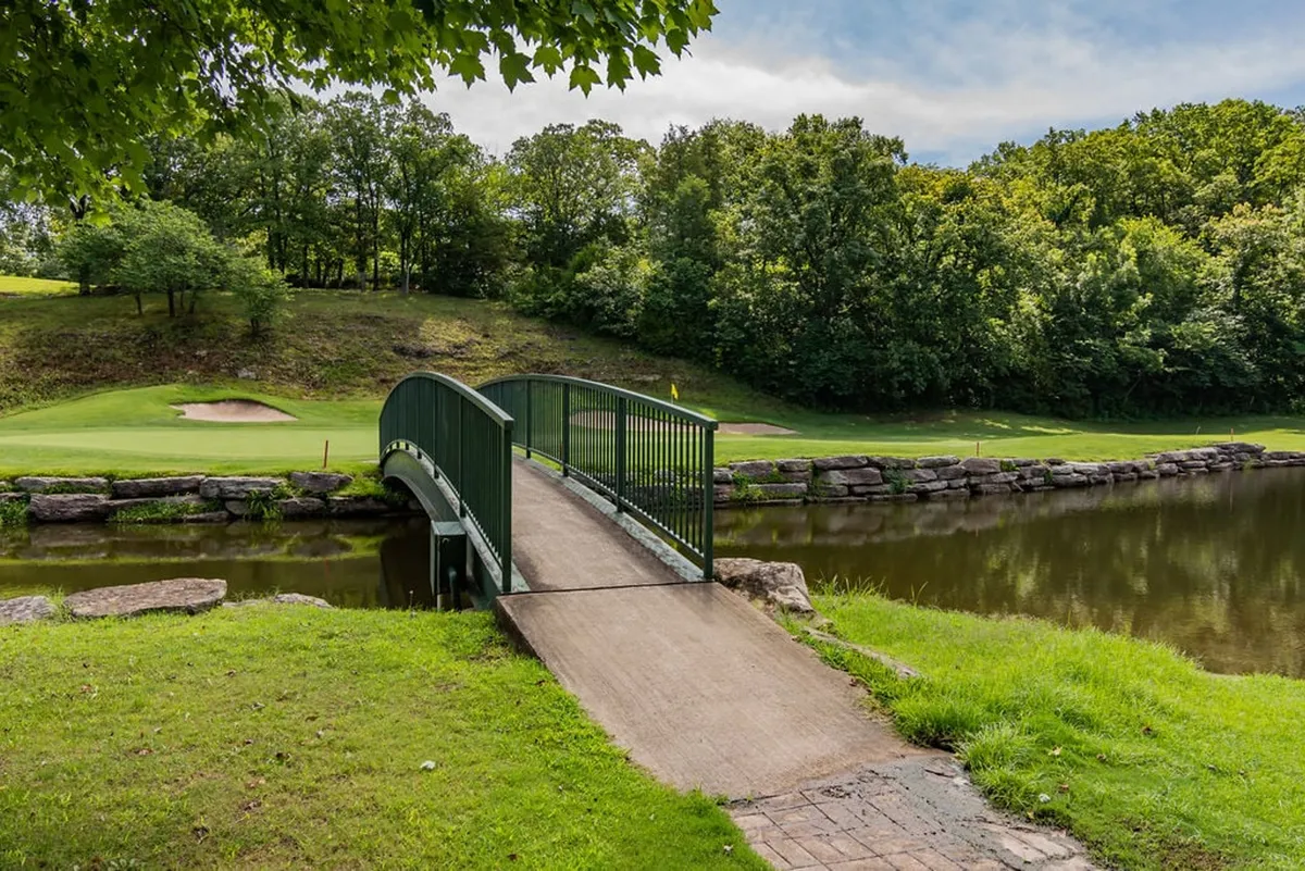 A tranquil exterior scene featuring a picturesque bridge over a pond with the golf course and trees in the background. Activities like walking, golfing, and relaxing by the water are implied, and the setting is peaceful and nature-rich.