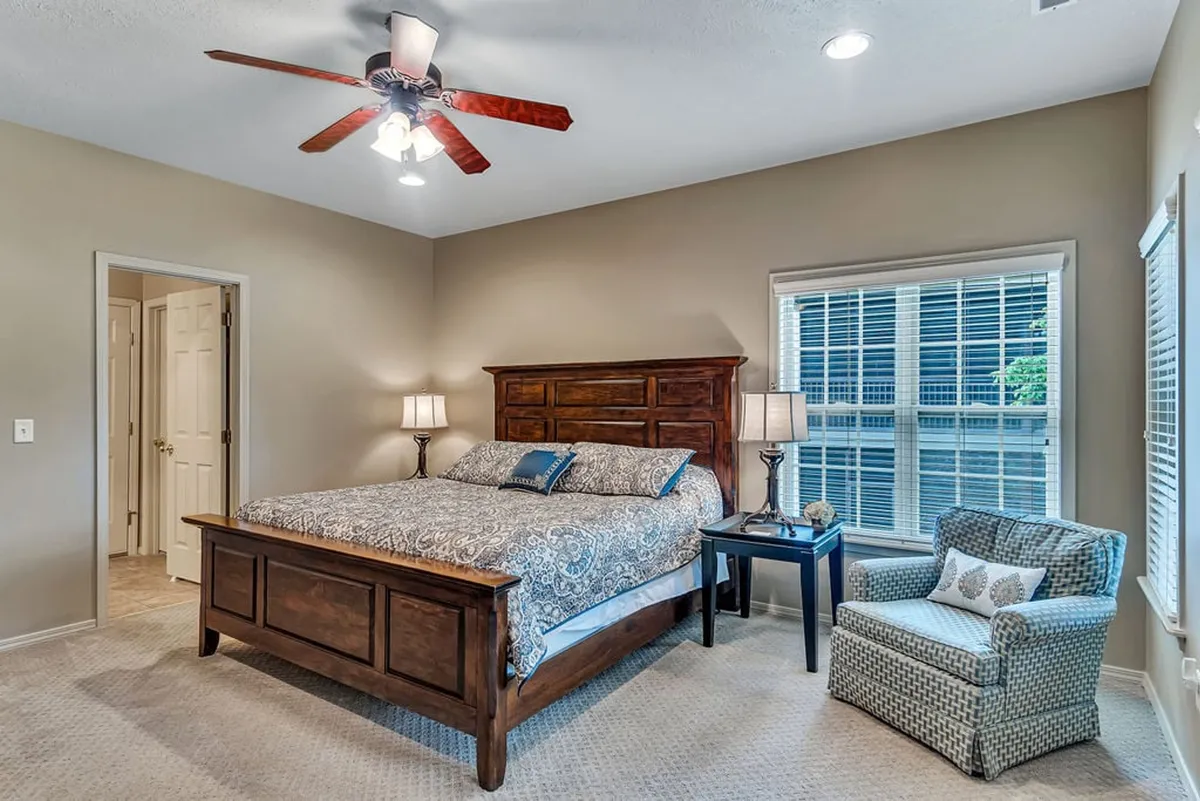 A serene bedroom featuring a large bed with plush bedding, flanked by nightstands and chairs, with sunlight streaming in through the window. The neutral palette and soft lighting evoke a tranquil, restful feel.