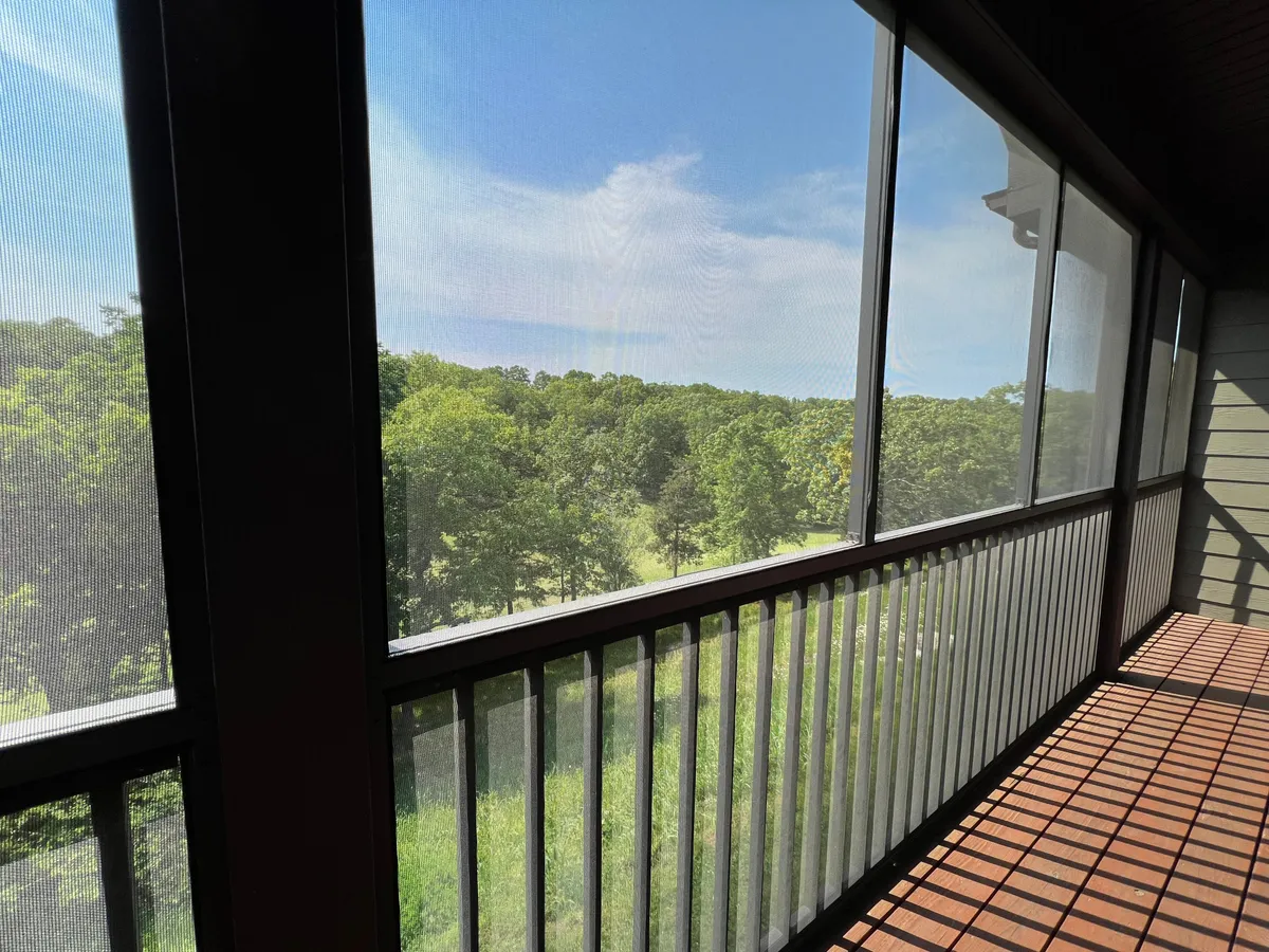 A spacious screened balcony with a table and chairs, surrounded by vibrant greenery. The abundance of natural light pours in, creating an inviting space for morning coffee, reading, or simply soaking up the views. The balcony overlooks the landscape, emphasizing the seamless blend of indoor and outdoor living.