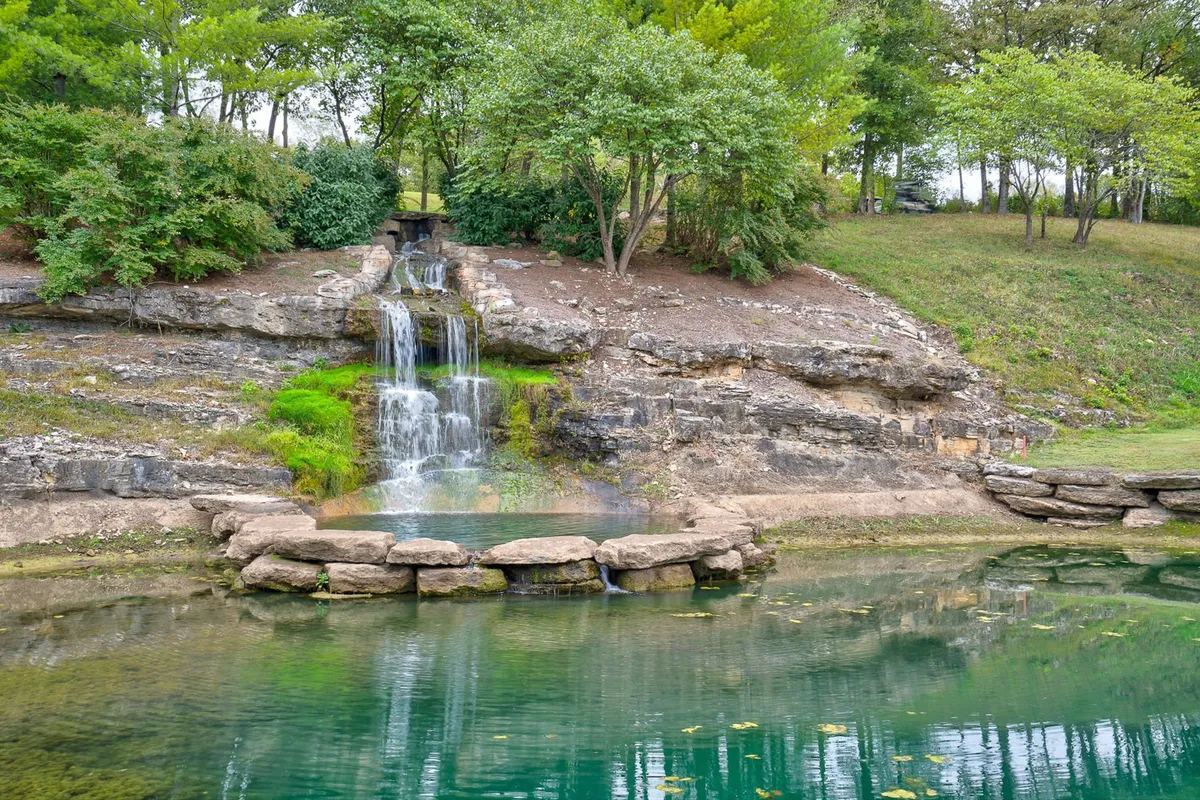 A lush, verdant scene featuring a picturesque waterfall cascading into a reflective pond, nestled among rock formations and surrounded by dense greenery. The area feels secluded and perfect for nature walks, birdwatching, or quiet moments spent observing local wildlife.