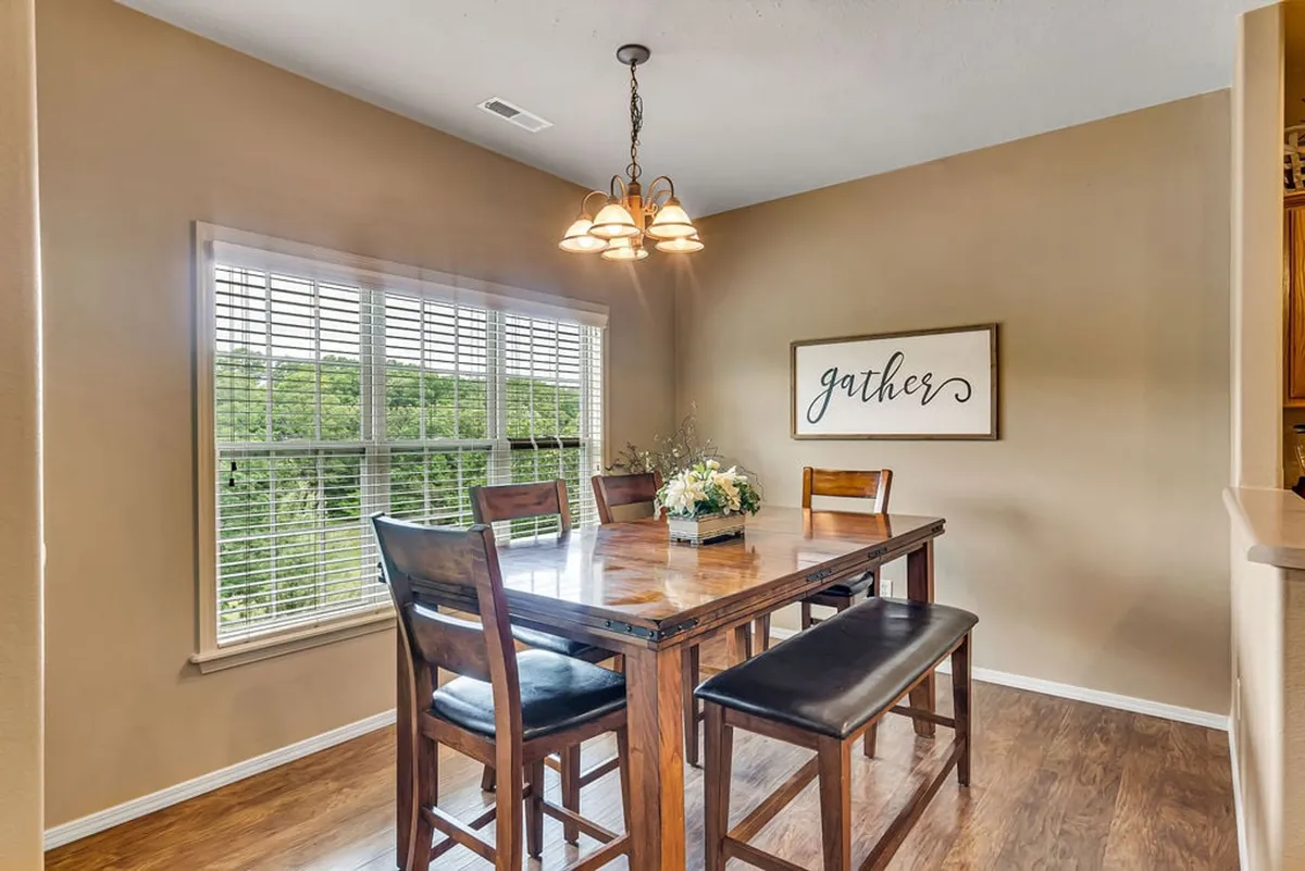 A warm dining area centered around a large wooden table, with four chairs and a bench beneath a chandelier. A decorative 'gather' sign and a sunny window view enhance the inviting, communal mood.
