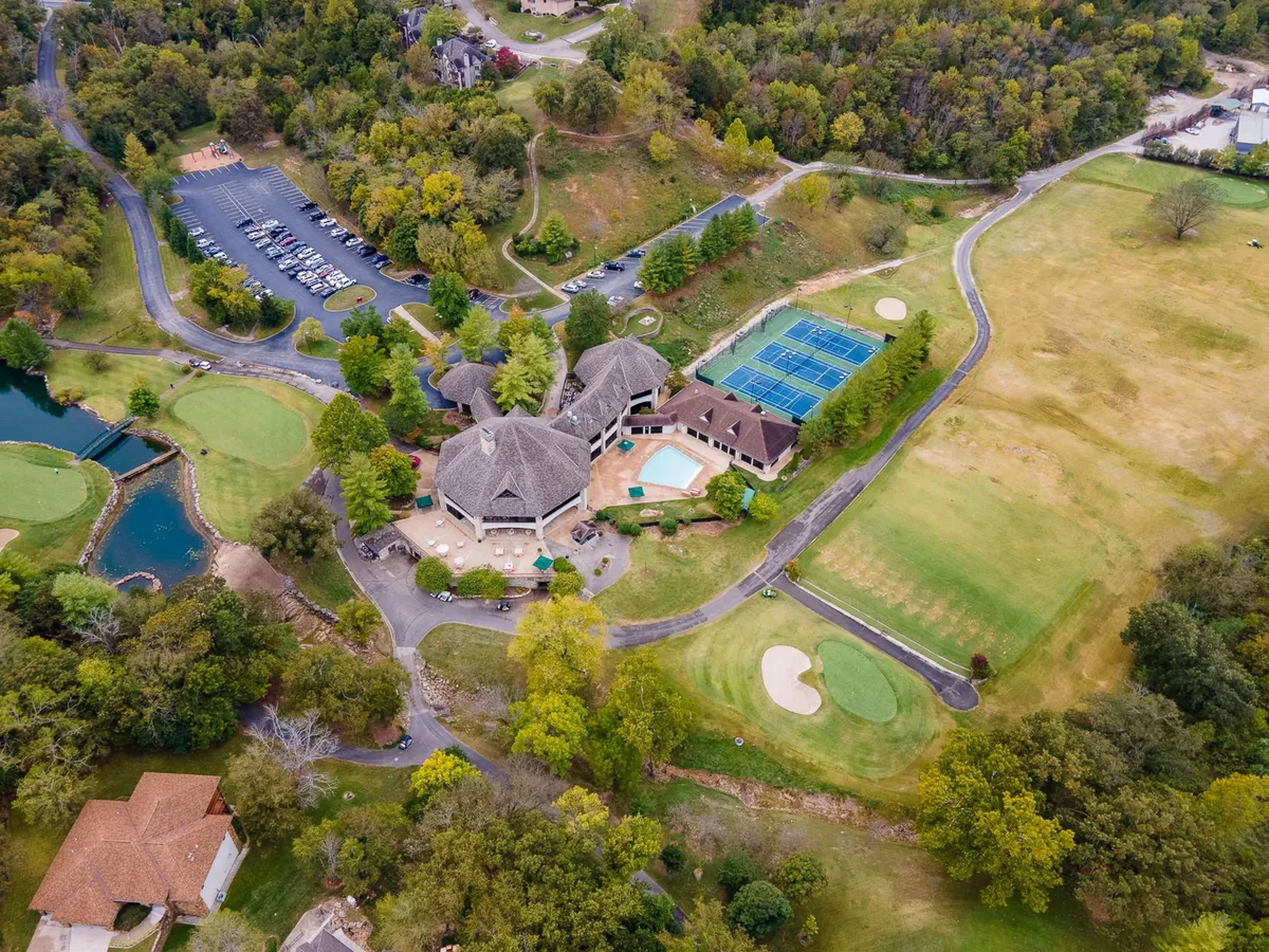 A vibrant exterior scene capturing the StoneBridge resort clubhouse surrounded by the golf course, tennis courts, a swimming pool, and parking area. The image provides a sweeping overview of multiple core amenities in a single frame: the championship golf course, the pool, and tennis facilities.